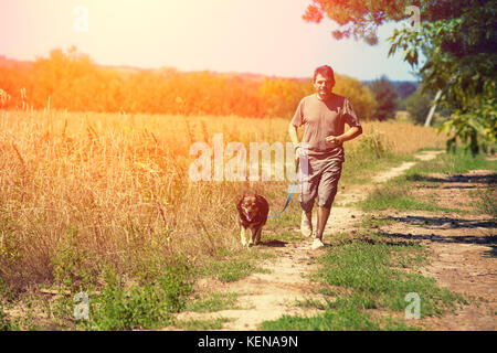 Un uomo con un cane al guinzaglio cammina lungo una strada di campagna nei pressi di un campo di grano in estate Foto Stock