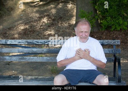 Closeup ritratto, vecchio uomo che impugnano il petto, aventi il dolore del cuore, seduti su una panchina, isolata all'esterno, verdi alberi sfondo. infarto del miocardio, aort Foto Stock