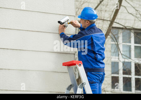 Maschio maturo tecnico permanente sulla scala del montaggio telecamera TVCC sulla parete Foto Stock