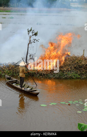 Donna asiatica sulla barca a remi, masterizzare albero secco, foglie essiccate al campo di pulizia nella stagione allagata, fiamma di fuoco su Causeway, fumo volare all'ambiente Foto Stock