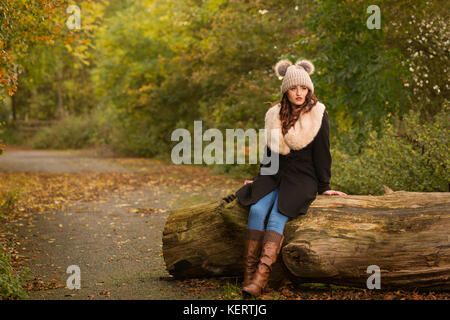 Donna vestita in cappotto e cappello di lana seduta sul log in autunno sunshine Foto Stock