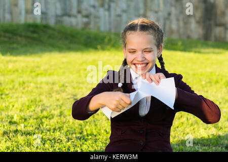 Ragazza in uniforme scolastica si siede su erba e strappa un pezzo di carta Foto Stock
