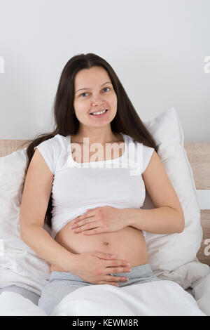 Felice donna incinta seduta sul letto di casa Foto Stock