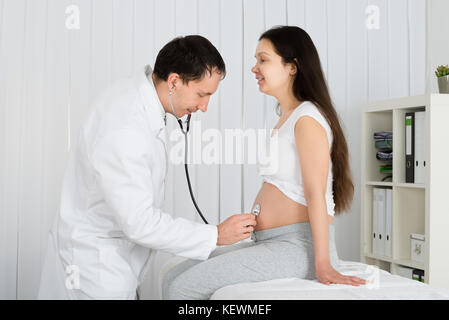 Medico utilizzando uno stetoscopio esaminando bianco donna incinta in clinica Foto Stock