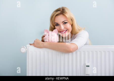 Giovane donna felice con piggybank sul radiatore a casa Foto Stock