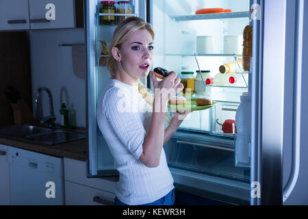 Giovane donna in piedi in cucina mangiare ciambella segretamente da frigo Foto Stock