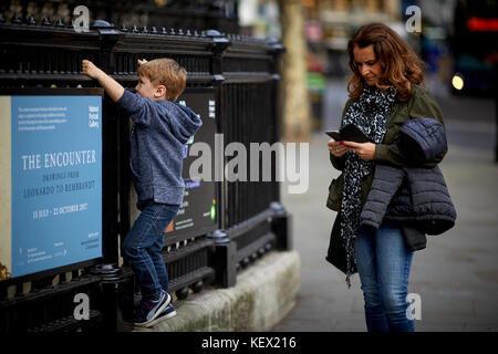 Mamma texting come bambino gioca sulle ringhiere alla National Portrait Gallery di Londra la città capitale d'Inghilterra Foto Stock