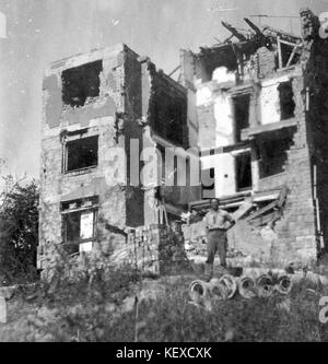 Una fotografia in bianco e nero che raffigura un edificio a più piani gravemente danneggiato, probabilmente a causa della distruzione in tempo di guerra, con un uomo in piedi davanti alle rovine. Foto Stock