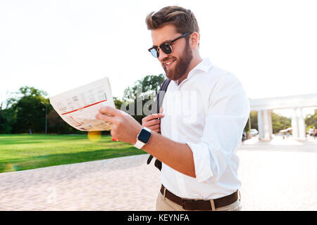 Sorridente uomo barbuto in occhiali da sole e vestiti business quotidiano di lettura all'aperto Foto Stock