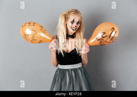 Crazy donna bionda in clown make-up holding palloncino di halloween e sorridente isolate su uno sfondo grigio Foto Stock