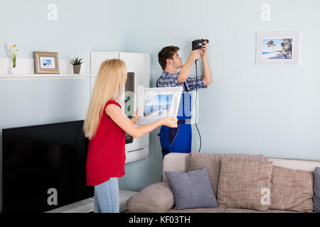 Donna che guarda la giovane utilizzando il trapano elettrico sul muro bianco a casa Foto Stock