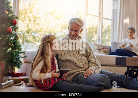 Bambina e nonno seduto sul pavimento da albero di natale con tavoletta digitale, con gran Madre seduti sul divano sul retro. Foto Stock