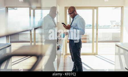 Uomo vestito in formali usando il telefono cellulare mentre faceva colazione a casa. uomo d'affari che controlla il telefono cellulare mentre beve un caffè in cucina. Foto Stock