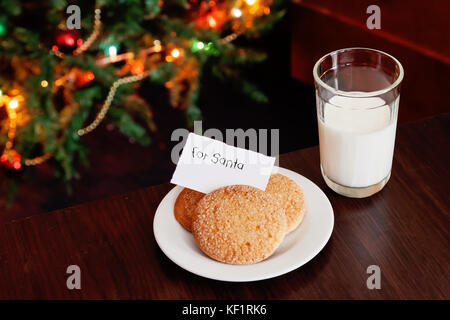 Biscotti di Natale e latte con nota per santa davanti a luci del nuovo anno albero. Foto Stock