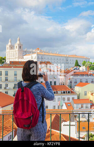 Viaggio di una giovane donna, vista posteriore di una turista solista che indossa uno zaino rosso scattando una foto degli edifici nel distretto di Alfama di Lisbona, Portogallo. Foto Stock