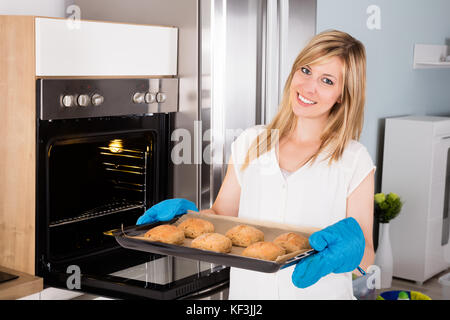 Giovane donna sorridente tenendo il vassoio pieno di Cookies vicino a un forno a microonde Foto Stock
