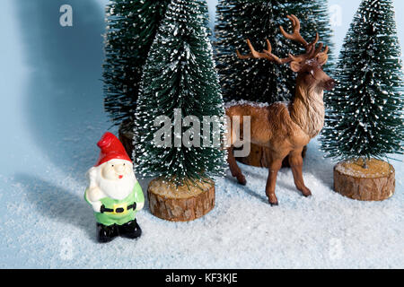 Allestimento completo di abeti artificiali come una piccola foresta innevata albero con una statuetta renne interno e un giardino gnome accanto su una vivace sfondo blu. Foto Stock