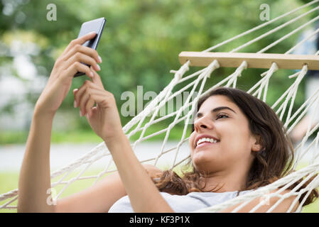 Giovane donna felice rilassante in amaca utilizzando il telefono cellulare in posizione di parcheggio Foto Stock