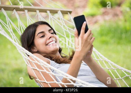 Giovane donna felice rilassante in amaca utilizzando il telefono cellulare in posizione di parcheggio Foto Stock