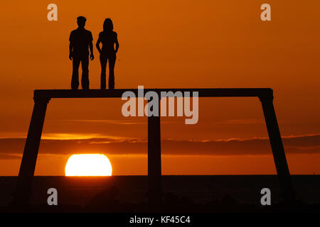 Il sole sorge sopra la scultura di coppia di Sean Henry sulla costa del Mare del Nord a Newbiggin Bay in Northumberland. Foto Stock