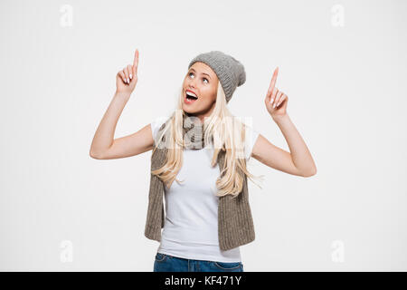 Ritratto di un felice Allegra donna in inverno cappello e sciarpa che puntano a due dito in alto isolato su sfondo bianco Foto Stock