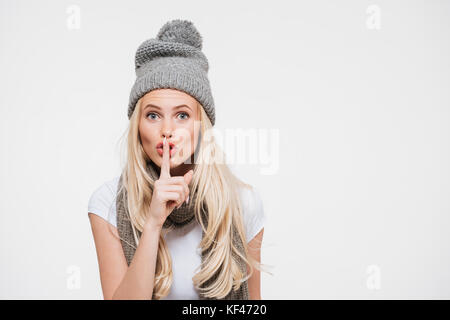 Ritratto di una giovane donna bionda in inverno cappello e sciarpa che mostra il silenzio gesto con il dito sulla sua labbra isolate su sfondo bianco Foto Stock