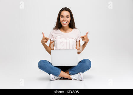 Ritratto di un felice giovane ragazza asiatica che puntano a due dita alla fotocamera mentre è seduto con computer portatile sul suo giro isolate su sfondo bianco Foto Stock