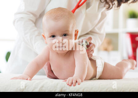 Pediatra esamina neonato ragazzo. medico utilizzando uno stetoscopio per ascoltare il bambino torna il controllo del battito cardiaco. kid sta guardando la telecamera. Foto Stock