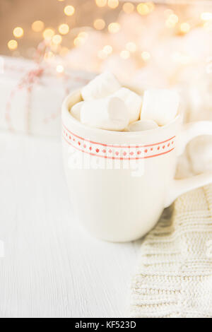 Tazza bianca con cioccolata calda bevanda di cacao e marshmallows sulla sommità. Vini spumanti ghirlanda di luci in background. felpa lavorata a maglia confezione regalo. tenui colori pastello Foto Stock