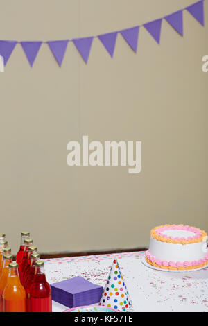 Bottiglie di rosso e succo di arancia accanto a viola di tovaglioli, un cono di partito hat e un bianco torta di compleanno in una sala decorata per la celebrazione Foto Stock