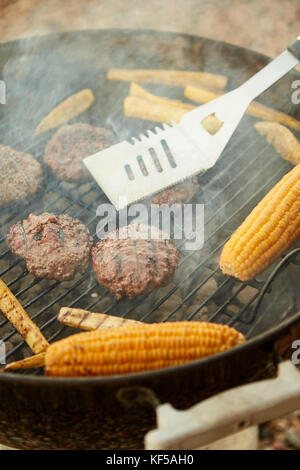 La cottura sulla pannocchia di mais e polpette di carne su un fuoco in un barbecue portatile utilizzando una spatola di metallo in una vista ravvicinata attraverso fumo Foto Stock