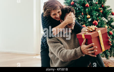 Giovane donna che copre gli occhi del suo partner durante la consegna di un regalo di Natale. Coppia giovane trascorrendo momenti felici mentre festeggia il Natale a casa. Foto Stock