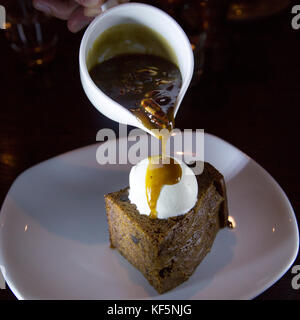 Sticky toffee pudding servito con gelato alla vaniglia. una brocca di salsa, cucita con noci pecan, viene versata oltre il tradizionale dessert britannico. Foto Stock