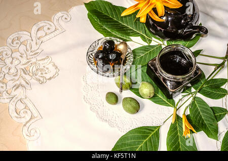 Inceppamento del noce in un vasetto di vetro con un nero teiera in porcellana con gigli arancione del noce ramoscelli giacente su una tovaglia ricamata Foto Stock