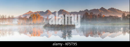 Ampio panorama cucita, autunno riflessione nel lago, misty sunrise. sun luce colpisce le alte montagne di picco. Foto Stock