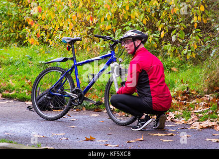 Un ciclista invecchiato in completo equipaggiamento da bicicletta fa alcune riparazioni in esecuzione alla sua bicicletta in una giornata umida in autunno Foto Stock