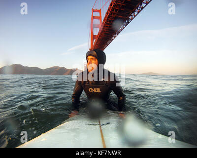 Stati Uniti, California, san francisco, fort point, surfer seduto su una tavola da surf sotto il Golden Gate bridge al tramonto Foto Stock