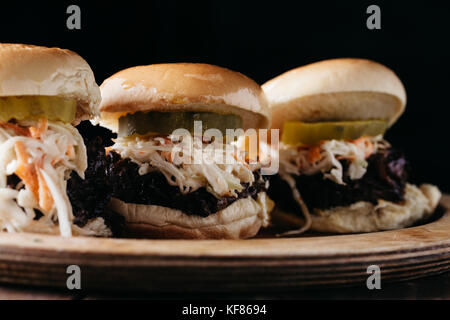 Cursori con estratto di carne di maiale, sottaceti e cole slaw su un piatto di legno con sfondo scuro Foto Stock