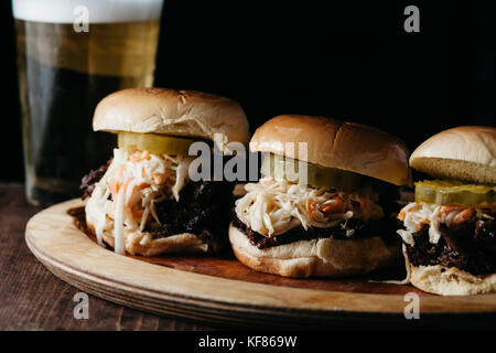 Cursori con estratto di carne di maiale, sottaceti e cole slaw su un piatto di legno con sfondo scuro Foto Stock