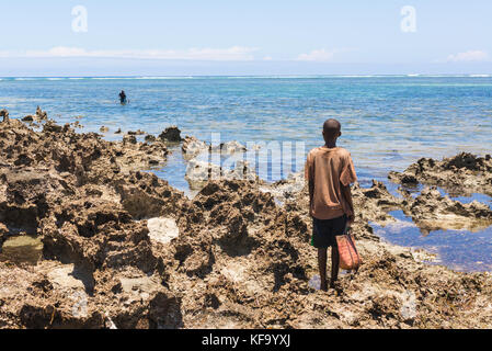 Giovane ragazzo africano che porta con sé una vecchia borsa della spesa e si trova su una costa rocciosa in attesa di un pescatore di lancia che stava tornando dal mare. Mare blu e. Foto Stock