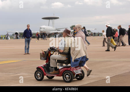 Uomo seduto in alimentato elettricamente con sedia a rotelle donna camminare a fianco con Royal Air Force RAF Boeing E-3D Sentry AEW-1 parcheggiato in static-dis Foto Stock
