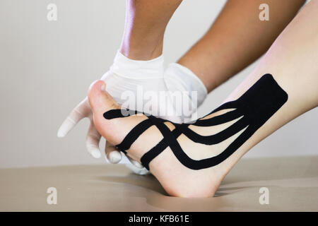 Medico fisioterapista con guanti applicazione medicale kinesio taping sulla caviglia sinistra di una paziente donna caucasica Foto Stock