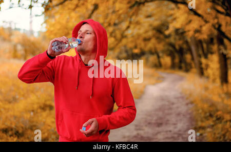 Atletica Giovane uomo in un rosso giacca sportiva con una cappa bere acqua dalla bottiglia dopo il jogging in un colorato Giallo autunno foresta. Foto Stock