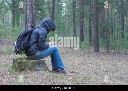 Una donna si siede su un moncone nel bosco di prima mattina e controlla il suo cellulare. Una femmina di escursionista, backpacker trekking nei boschi e montagne. H Foto Stock