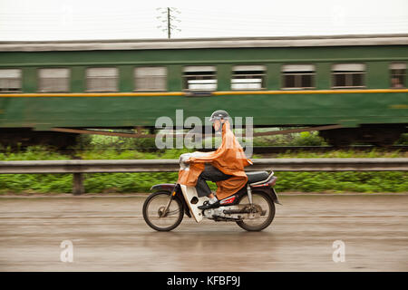 Il Vietnam, Hanoi, campagna, un uomo passa un treno sul suo ciclomotore sotto la pioggia Foto Stock