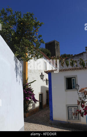 Il cielo blu e un albero di limone sovrastante le stradine acciottolate della città medievale di Obidos, Portogallo. La torre del castello in background. Foto Stock