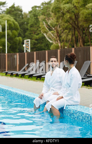 Giovani coppie sedute vicino alla piscina con gambe in acqua e chiacchierate al resort Foto Stock