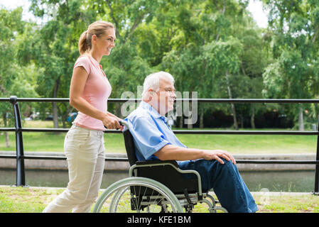 Vista laterale di un sorridente giovane donna che assiste il suo padre disabili su sedia a rotelle in posizione di parcheggio Foto Stock