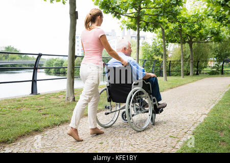 Vista laterale di un sorridente giovane donna che assiste il suo padre disabili su sedia a rotelle in posizione di parcheggio Foto Stock