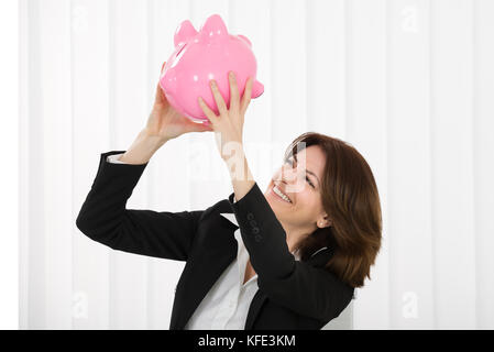 Felice imprenditrice scuotendo piggybank indossando occhiali in ufficio Foto Stock